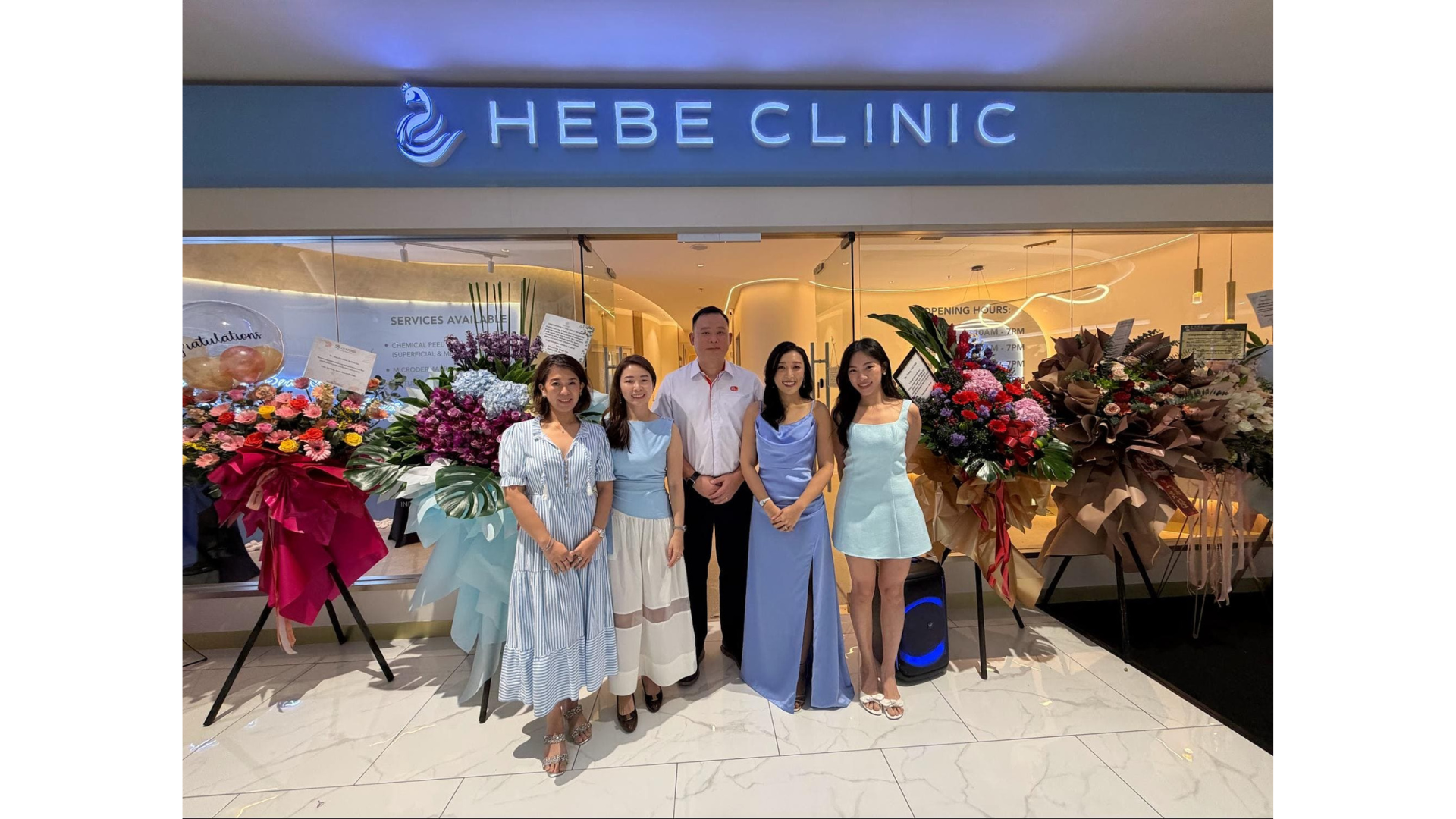 Group photo of Hebe Clinic team members standing outside the storefront, surrounded by congratulatory flower bouquets to mark the clinic's 1st anniversary and grand opening of the KLGCC branch