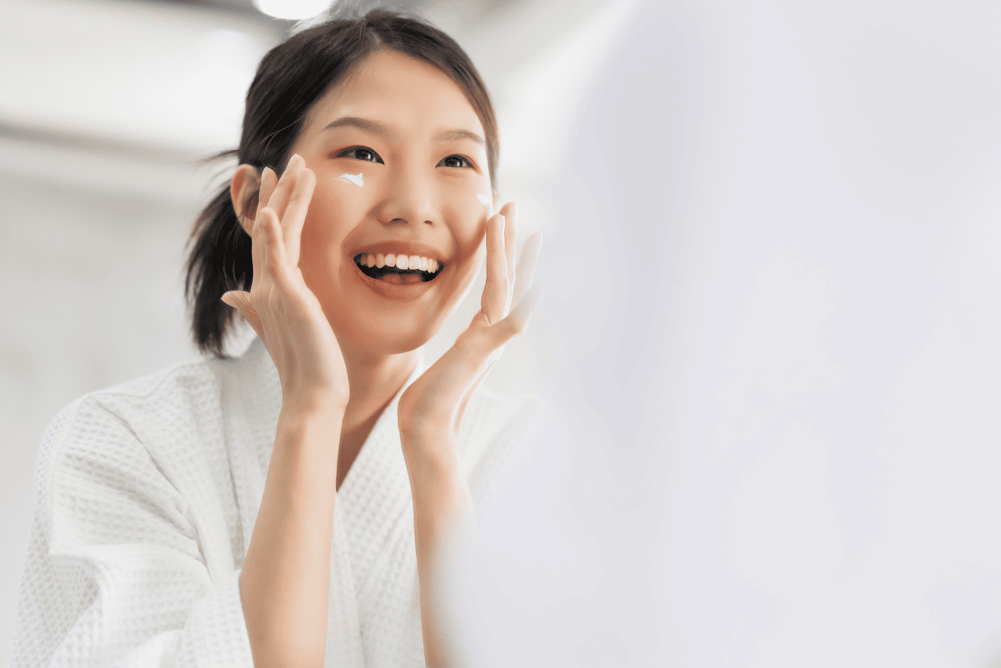 Smiling woman in a white robe applying face cream to her cheeks during her skincare routine