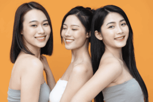 Three smiling women with bare shoulders pose together against an orange background, showcasing smooth, healthy skin