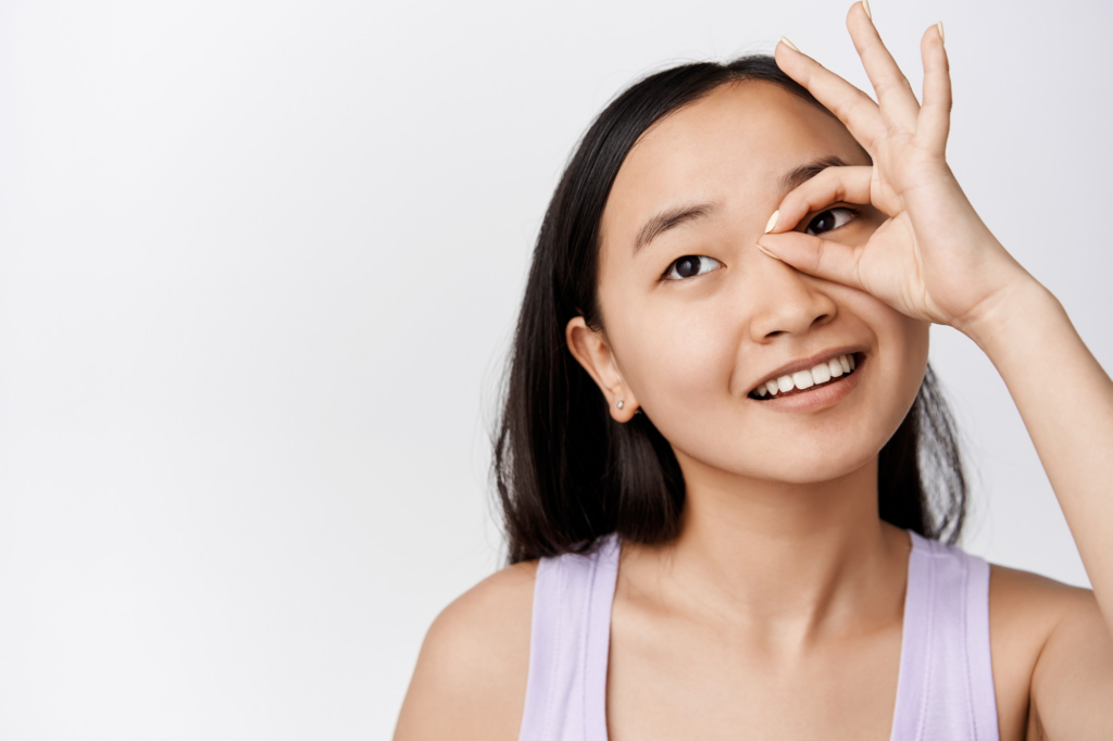 A smiling woman playfully frames her eye with her fingers in an "OK" gesture