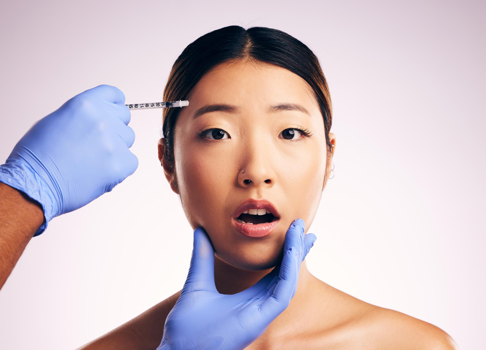 A woman looks at the camera with a surprised expression as a gloved hand holds a syringe near her forehead and another hand touches her chin