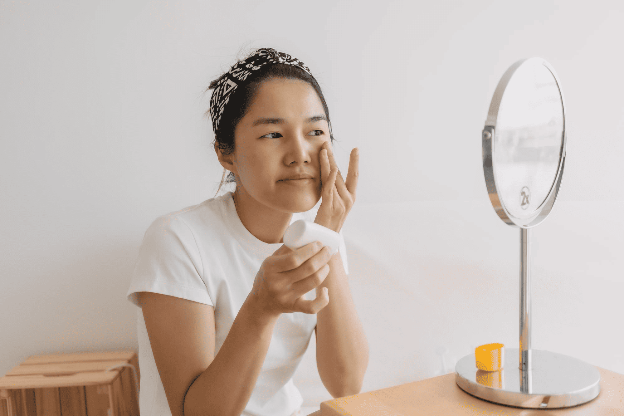 A woman applies skincare cream to her face while looking in a small standing mirror