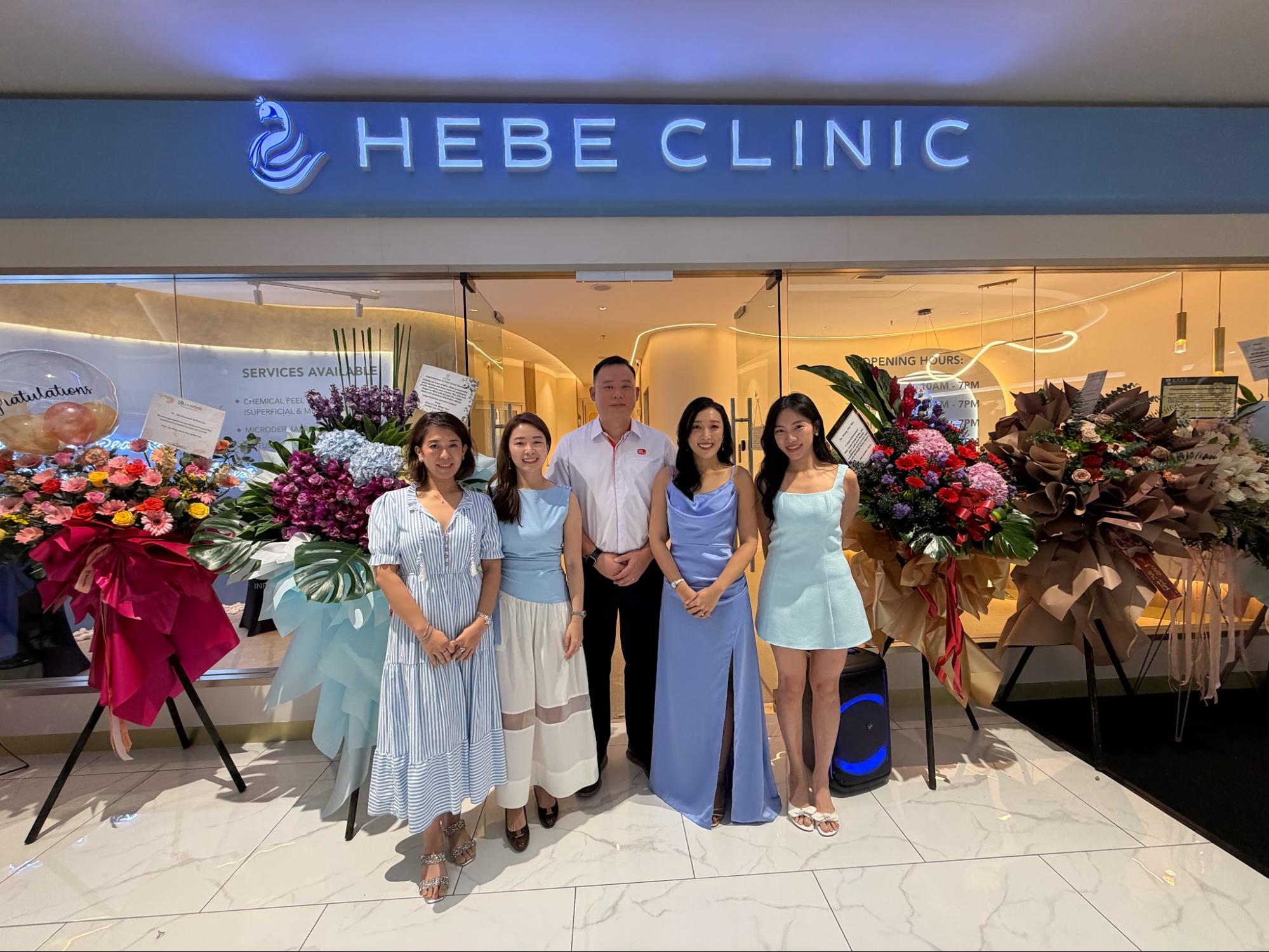 Group photo of Hebe Clinic team members standing outside the storefront, surrounded by congratulatory flower bouquets to mark the clinic's 1st anniversary and grand opening of the KLGCC branch