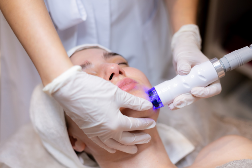 A woman receives a facial skin treatment as a technician in gloves applies a light-based device near her jawline.